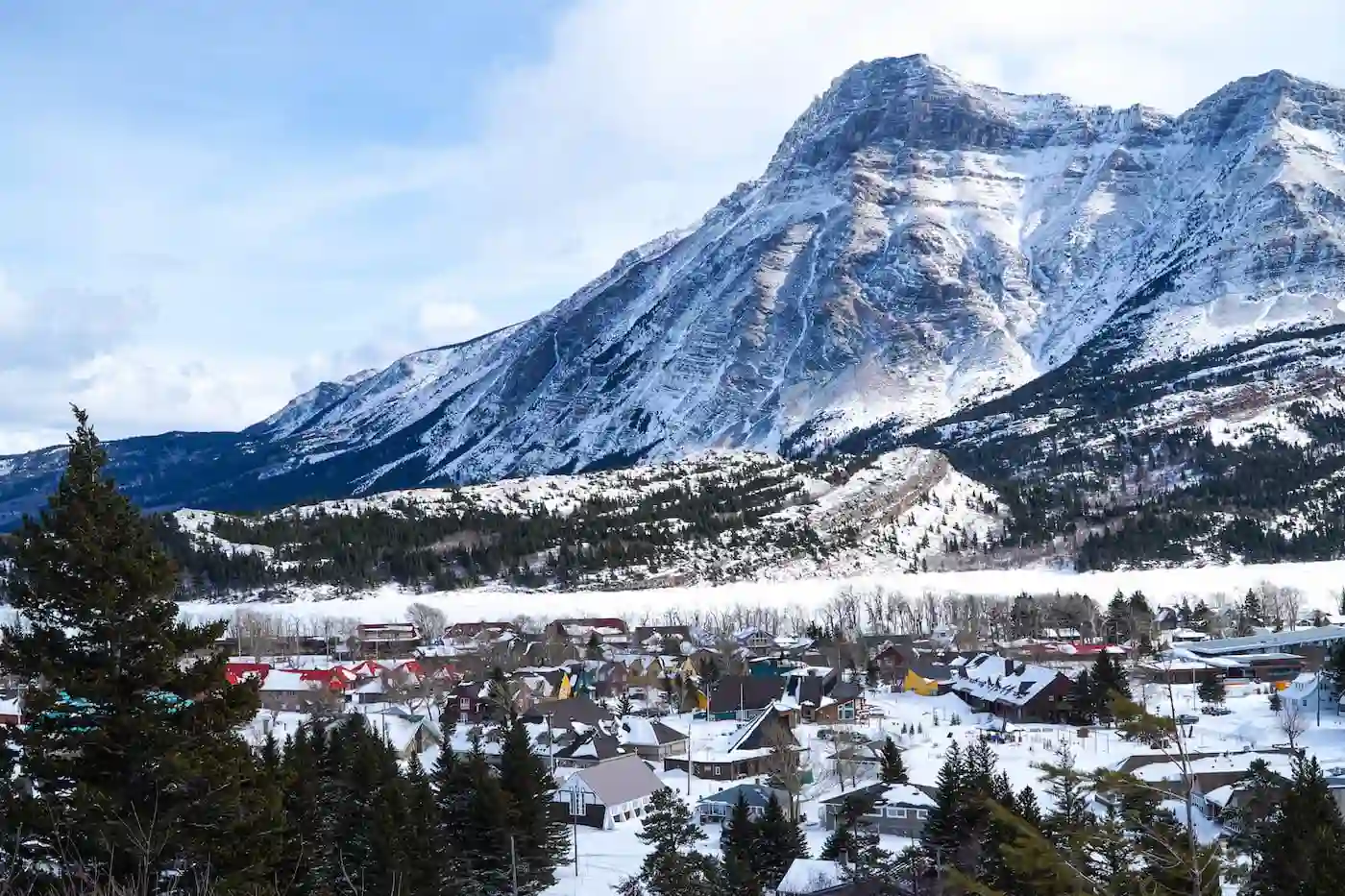 Dramatic and photogenic of a mountain in waterton national park with the frozen lake in the middle and slightly overlooking the townsite from a trail of trees framed by evergreens.
