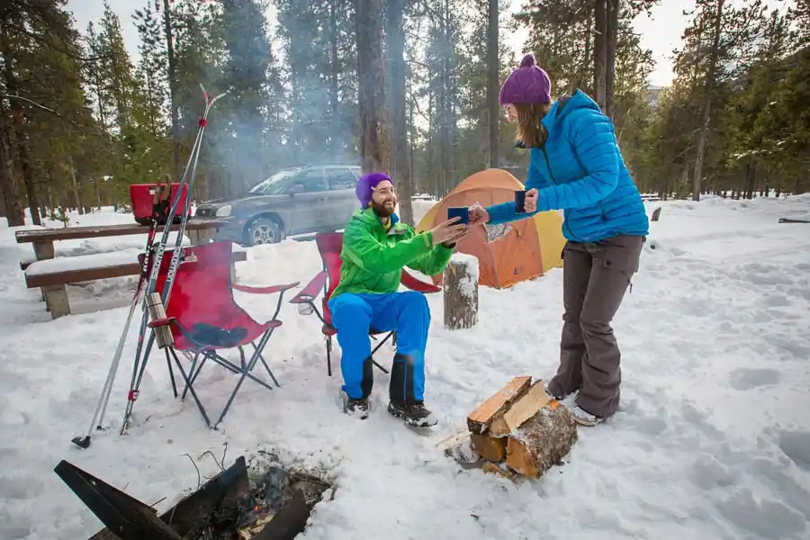 Image of man and woman in winter clothes camping in the winter in Waterton National Park sharing a hot drink with firewood in front of an active fire pit and a cat in the background in the middle of the forest.